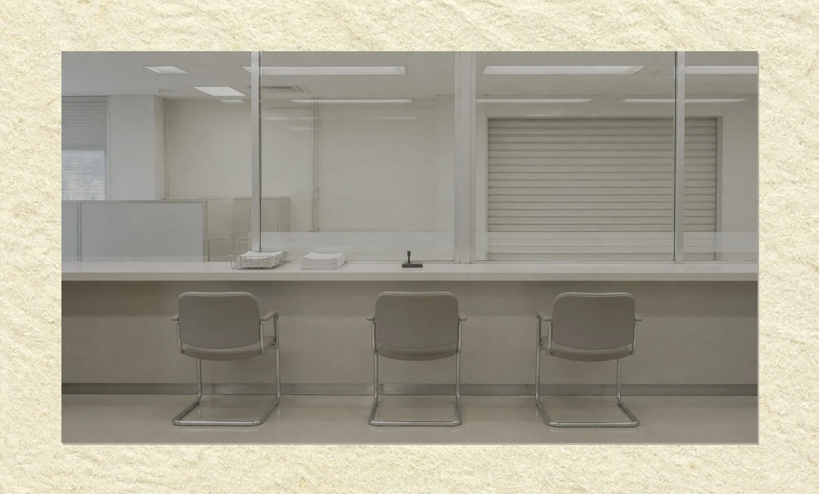 An empty public service counter in a plain government office, photographed straight on. A long counter runs across the frame beneath a glass partition, with three unoccupied chairs in front of it, a tray of blank forms and a stamp holder on the surface, and a closed shutter at one window position.