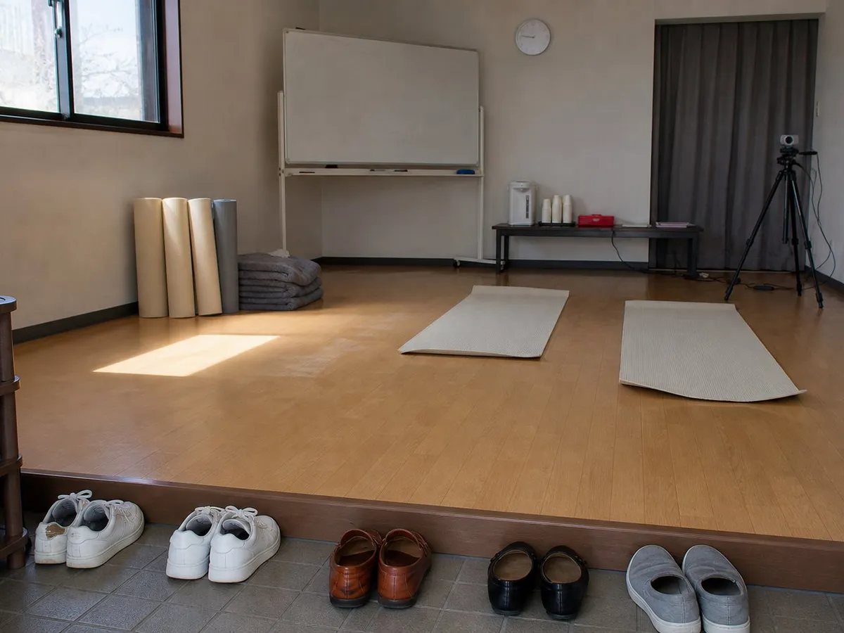 A photograph of a small rented room used for a yoga class, seen from the entrance step where five pairs of shoes are lined up: white sneakers, brown loafers, black flats and grey slip-ons. Inside, pale laminate flooring, two cream yoga mats still unrolled, rolled mats and folded blankets against the wall, a wiped whiteboard on a stand, a low table with a kettle, paper cups and a small red first-aid kit, a closed curtain across an alcove at the back, and a webcam on a tripod pointing at the empty floor. Late afternoon light falls from a window on the left. Nobody is present. AI-generated photo illustration; not a photograph of any real room, event or person.