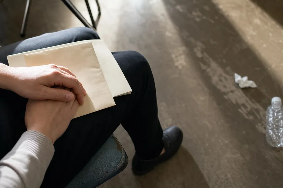 A close photograph taken from just above and behind a seated person, showing only their lap and their hands, so that no face and no upper body appear in the frame. A pale cream envelope and a document wallet rest across dark trousers, and both hands are folded on top of them with the fingers pressed together and slightly tense. The sleeves of a cream knitted jumper cover the wrists. Light falls from the left across the backs of the hands and the envelope, leaving the right side of the lap in shadow. Below and beyond the knees the floor of a waiting area recedes out of focus: a scuffed pale linoleum surface with the metal legs of other chairs, a crumpled tissue and a half-empty plastic bottle lying near them, and a broad band of window light across the middle distance.