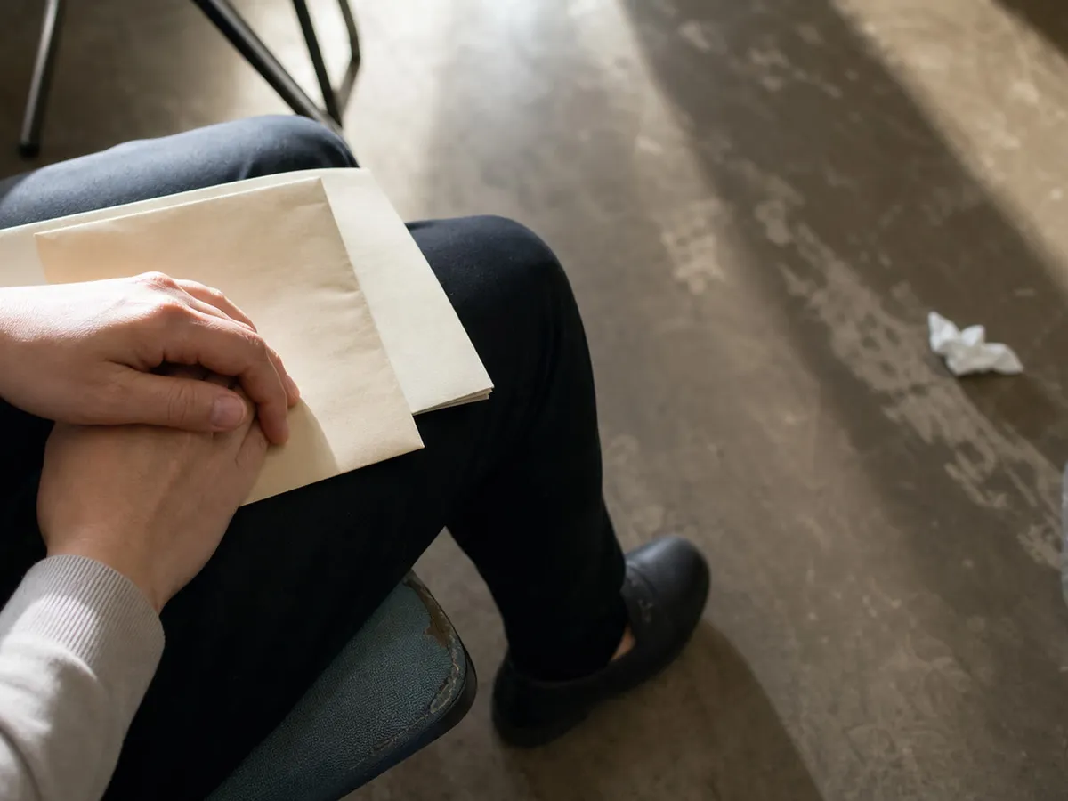 A close photograph taken from just above and behind a seated person, showing only their lap and their hands, so that no face and no upper body appear in the frame. A pale cream envelope and a document wallet rest across dark trousers, and both hands are folded on top of them with the fingers pressed together and slightly tense. The sleeves of a cream knitted jumper cover the wrists. Light falls from the left across the backs of the hands and the envelope, leaving the right side of the lap in shadow. Below and beyond the knees the floor of a waiting area recedes out of focus: a scuffed pale linoleum surface with the metal legs of other chairs, a crumpled tissue and a half-empty plastic bottle lying near them, and a broad band of window light across the middle distance.