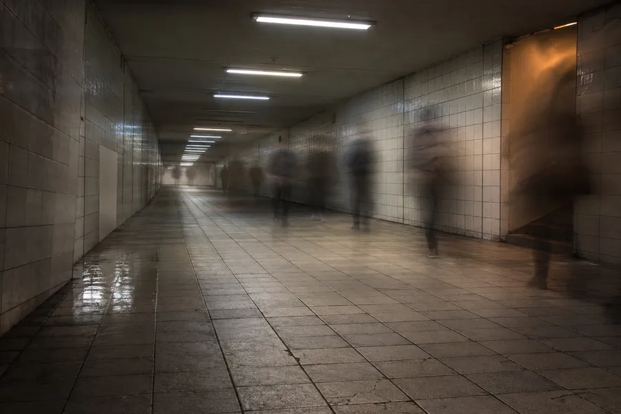 A wide pedestrian underpass in a Japanese railway station in the early evening, photographed on a long exposure so that the people walking through it appear only as soft, half-transparent streaks. No individual figure is sharp and no face, clothing detail or bag can be identified. The tiled floor is scuffed and slightly uneven, with a shallow puddle reflecting light near the left wall. A run of fluorescent strips crosses the ceiling, one of them dimmer than the rest. The walls are plain tile with one repaired panel in a mismatched shade, and there is no signage, advertisement or lettering of any kind anywhere in the frame. Strong overhead light falls away toward the far end of the passage, which is almost dark, and a single warm reflection from a stairwell out of frame on the right lands on the wall.