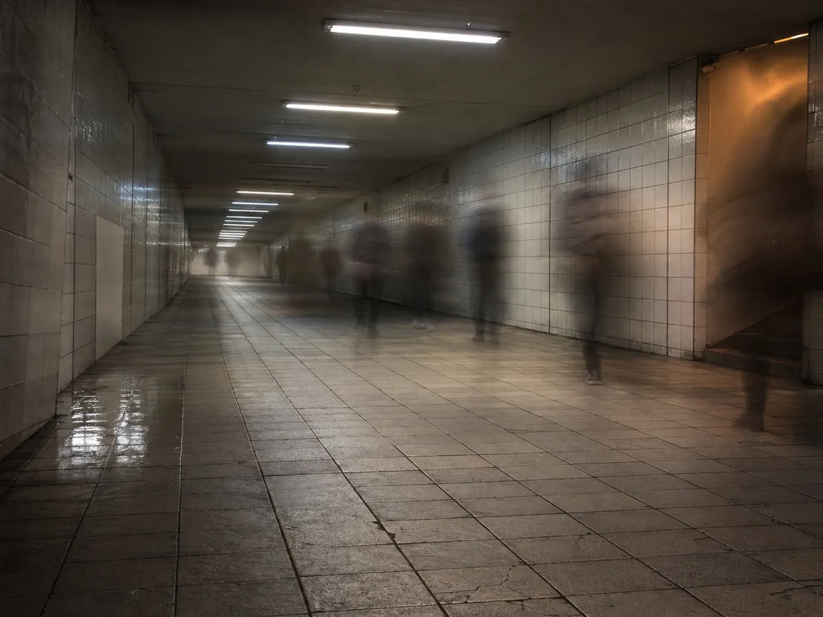 A wide pedestrian underpass in a Japanese railway station in the early evening, photographed on a long exposure so that the people walking through it appear only as soft, half-transparent streaks. No individual figure is sharp and no face, clothing detail or bag can be identified. The tiled floor is scuffed and slightly uneven, with a shallow puddle reflecting light near the left wall. A run of fluorescent strips crosses the ceiling, one of them dimmer than the rest. The walls are plain tile with one repaired panel in a mismatched shade, and there is no signage, advertisement or lettering of any kind anywhere in the frame. Strong overhead light falls away toward the far end of the passage, which is almost dark, and a single warm reflection from a stairwell out of frame on the right lands on the wall.