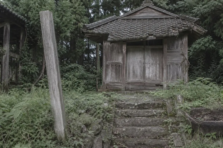 A photograph of a very small disused religious hall in the Japanese countryside in flat afternoon light. A short flight of worn stone steps, overgrown with tall summer weeds, leads up to a modest wooden building with a low tiled roof and closed weathered doors. A wooden gatepost leans out of true, a cracked stone basin holds dead leaves, the paint has gone from the timber and one roof tile has slipped. Cedar trees crowd in behind. Nobody is present and nothing suggests recent use. AI-generated photo illustration; not a photograph of any real building or organisation.
