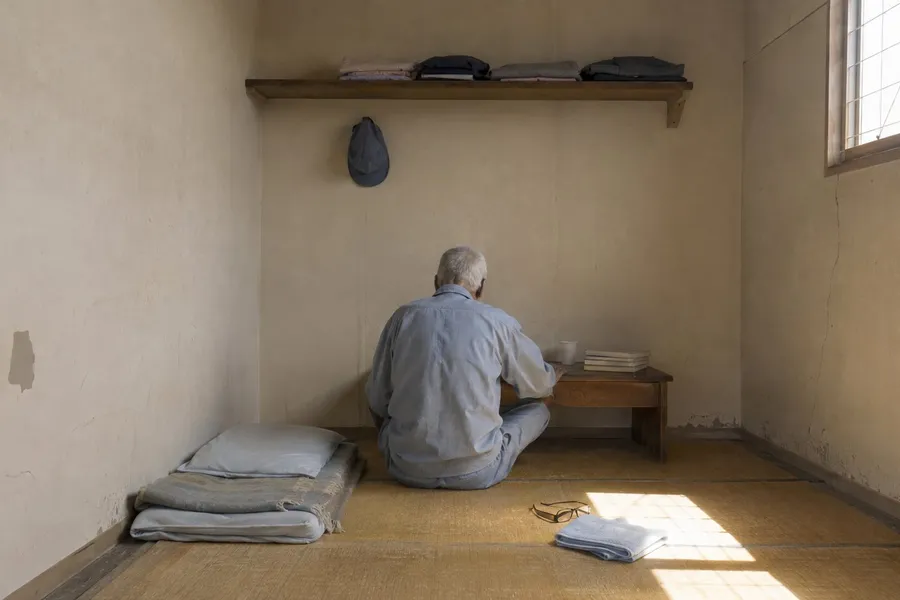 A small single-occupancy room in a plain institution, photographed from the doorway in the late afternoon. An elderly man sits on the floor with his back to the camera, facing a low wooden desk against the far wall. He wears an unmarked light grey-blue cotton work uniform, his back stooped and his hair short and white, and his face is entirely out of view. On the desk are a plastic cup and a few paperbacks laid flat so that no title can be read. A shelf above the desk holds a folded change of clothes, and a cloth cap hangs from a hook on the wall. A futon and a thin blanket are folded neatly on the tatami to the left. A window set high on the right wall, above head height and divided by thin metal glazing bars, throws a bright rectangle of low daylight across the matting, where a pair of reading glasses and a folded towel lie on the floor. One panel of the tatami is worn paler than the rest, in the place where someone habitually sits. Nobody else is present.