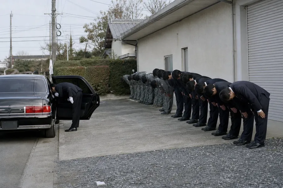 A photograph of a concrete driveway beside a plain low building on a bright cold morning. A long black chauffeur-driven sedan waits at the kerb with its rear door held open by a driver in a dark suit and white cotton gloves, who is bowing. Along the wall a line of about twelve men bows deeply toward the car in unison, each bent sharply from the waist with the head dropped and hands pressed to the thighs, so that no face is visible; those nearest the car wear dark double-breasted suits and those behind them wear grey work jackets. A rolled steel shutter, a clipped hedge, grey utility poles with bundled cables and gravel at the edge of the concrete. AI-generated photo illustration; not a photograph of any real place, organisation or person.