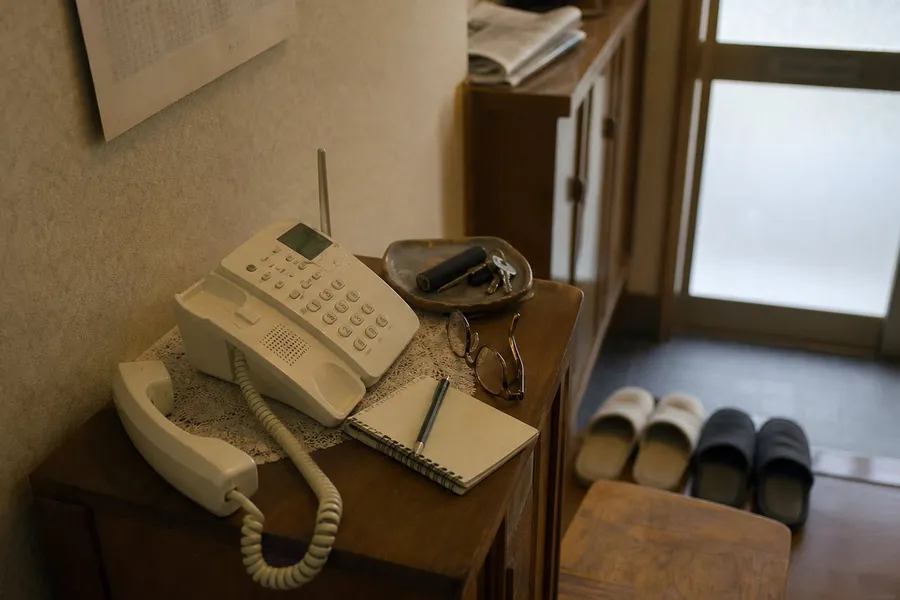 A photograph of the entrance hall of a Japanese house in the late afternoon, empty of people. On a low wooden telephone stand sits a cream-coloured cordless telephone with its handset lifted out of the cradle and lying beside it, its coiled cord hanging over the edge of the stand. Around the telephone are a spiral notepad with a ballpoint pen laid across it, a pair of reading glasses folded closed, a shallow dish holding keys and a cylindrical seal case, and a lace mat. Behind the stand a shoe cupboard carries a folded newspaper, slippers are arranged in pairs on the step up into the house, and a frosted glass front door lets in flat grey daylight. No people are present and no text is legible anywhere. AI-generated photo illustration; not a photograph of any real house, case or person.