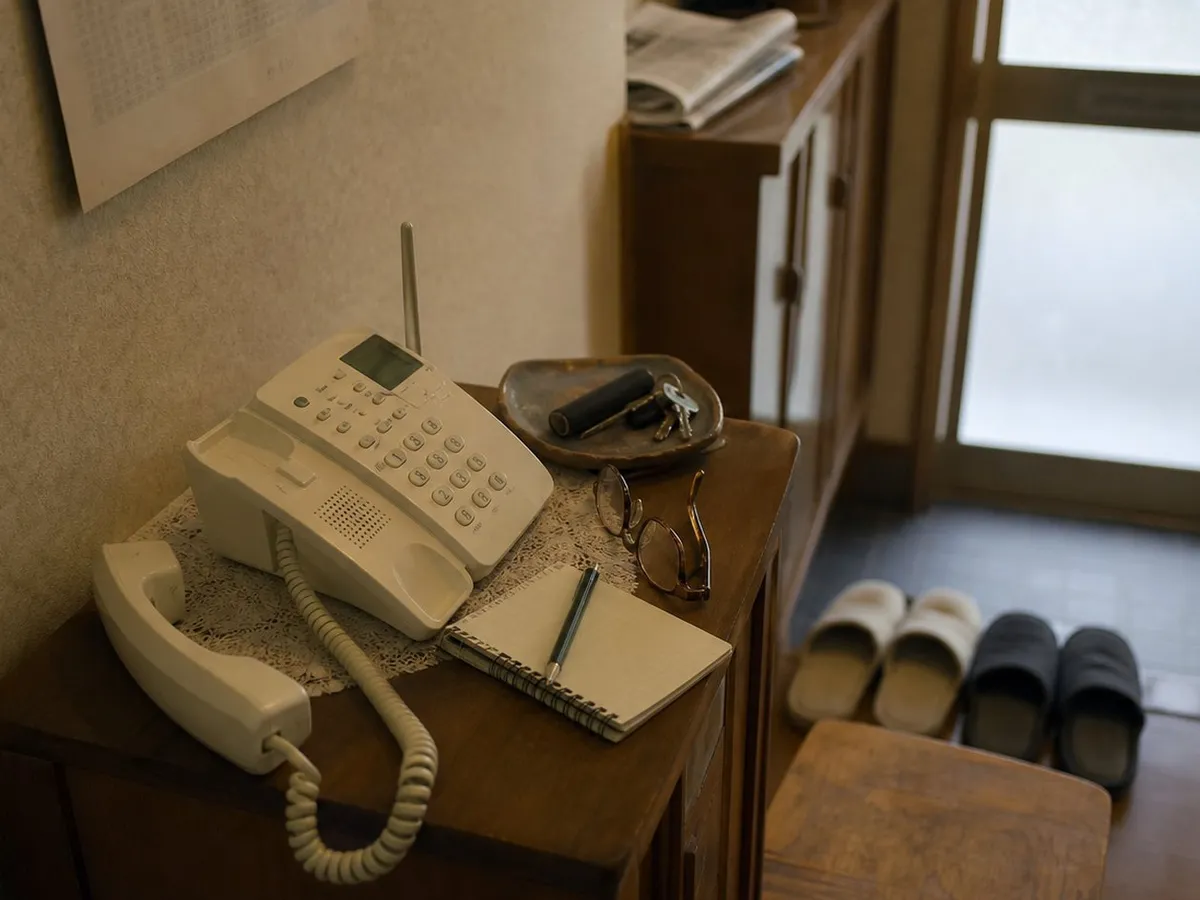 A photograph of the entrance hall of a Japanese house in the late afternoon, empty of people. On a low wooden telephone stand sits a cream-coloured cordless telephone with its handset lifted out of the cradle and lying beside it, its coiled cord hanging over the edge of the stand. Around the telephone are a spiral notepad with a ballpoint pen laid across it, a pair of reading glasses folded closed, a shallow dish holding keys and a cylindrical seal case, and a lace mat. Behind the stand a shoe cupboard carries a folded newspaper, slippers are arranged in pairs on the step up into the house, and a frosted glass front door lets in flat grey daylight. No people are present and no text is legible anywhere. AI-generated photo illustration; not a photograph of any real house, case or person.