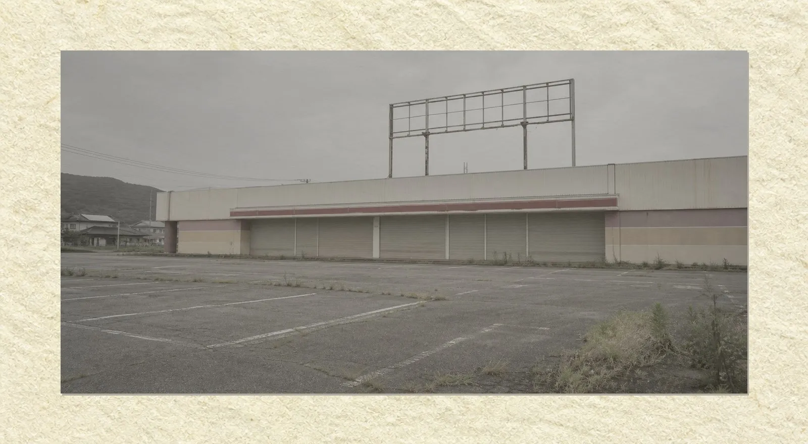 A closed-down pachinko parlour seen across an empty asphalt car park on an overcast afternoon. Metal shutters are rolled down across the whole entrance, an empty steel sign frame stands on the roof with its panel removed, and weeds grow through cracks in the parking area. Nobody is present.