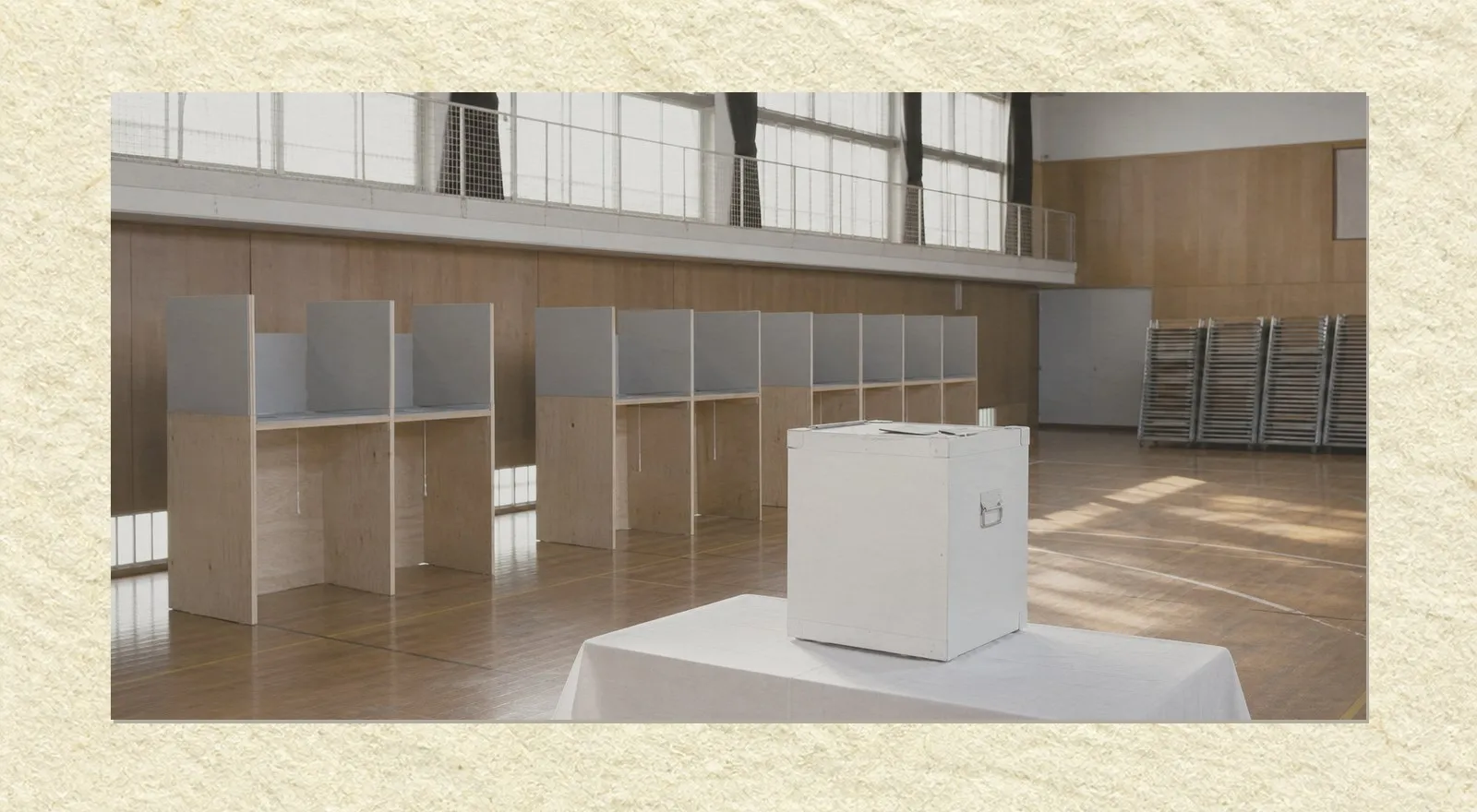 An empty polling station in a school gymnasium in the late afternoon. A row of plywood voting booths with grey privacy screens stands along a wooden wall, and a white ballot box sits on a cloth-covered table in the foreground. Stacked folding chairs stand against the wall at the right. Nobody is present.