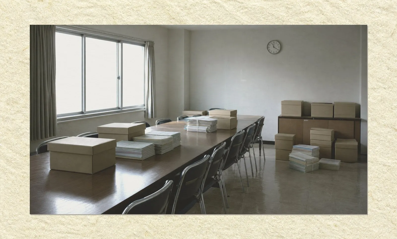 A muted photograph mounted on pale paper with a narrow margin. Inside a plain institutional meeting room, a long dark table runs from the foreground into the middle distance, ringed by empty steel-framed chairs pushed in. On the table and on the linoleum floor beside it stand unlabelled cardboard document boxes and thick bundles of paper files. Cool daylight enters through a frosted window on the left; the ceiling lights are off, and a plain round clock hangs on the far wall. No people are present. AI-generated photo illustration; not a photograph of any real room or investigation.