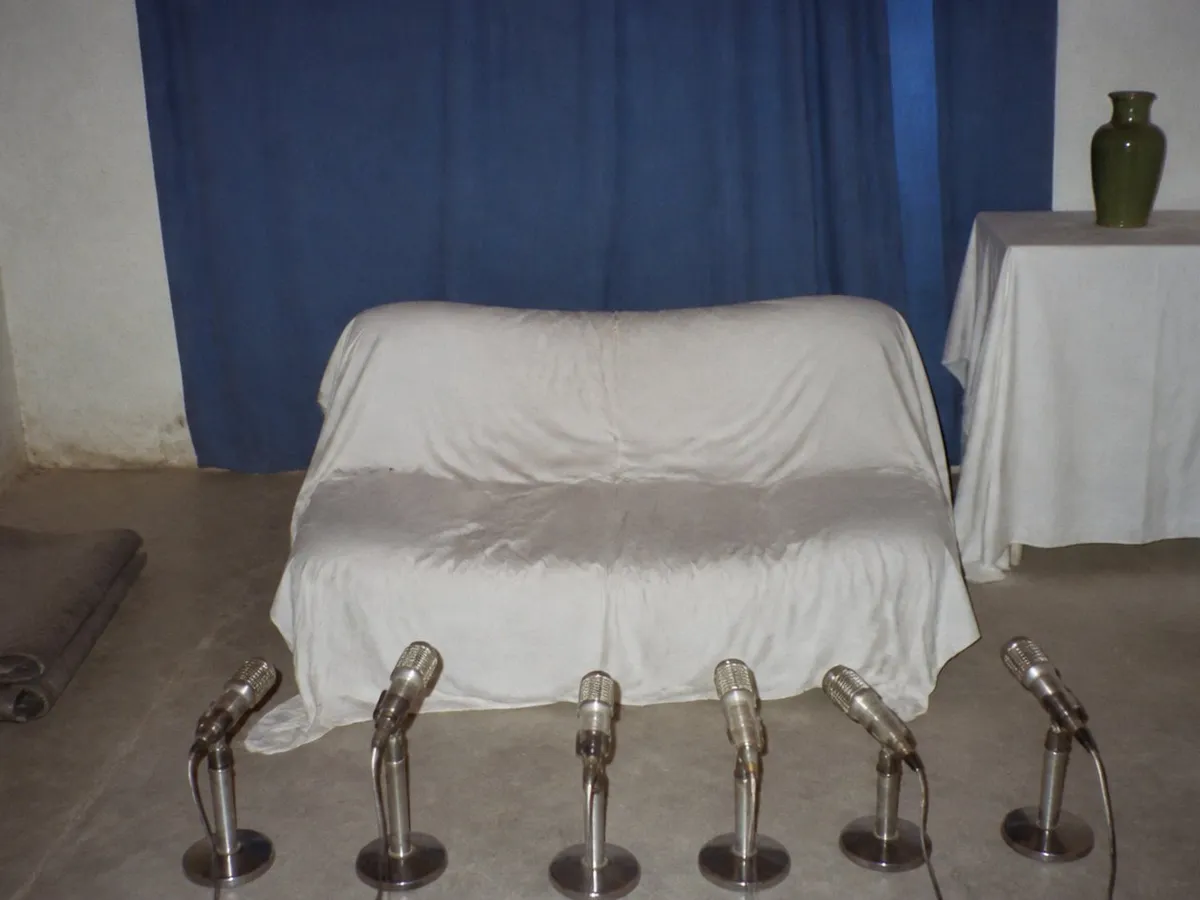 A photograph of a small plain room lit by direct camera flash, in the washed-out style of a 1990s amateur snapshot. At the centre a low couch draped in white cloth stands empty, the cloth still creased and dented where somebody had been sitting. Facing the empty seat, a row of six old silver microphones on short stands is lined up along the bare floor. A plain blue cloth curtain hangs slightly crooked on the wall behind, a low white-clothed table at one side holds a green ceramic vase, and a folded grey blanket lies on the floor. Nobody is present. AI-generated photo illustration; not a photograph of any real room, event or person.