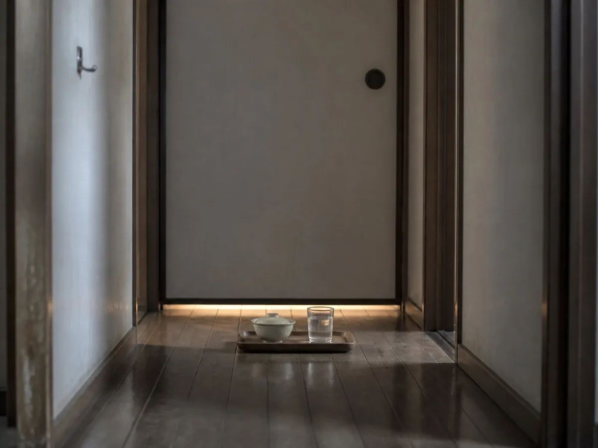 A photograph looking down a dim domestic hallway in an ordinary Japanese home. At the end of the hallway a plain sliding door is closed, with a thin line of light showing along its lower edge. On the floor immediately outside the door sits a small wooden tray holding a covered bowl and a glass of water, placed and untouched. The hallway is bare: a worn wooden floor, plain walls, a coat hook with nothing on it. No people are present. AI-generated photo illustration; not a photograph of any real home.