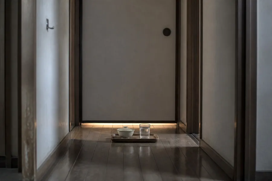 A photograph looking down a dim domestic hallway in an ordinary Japanese home. At the end of the hallway a plain sliding door is closed, with a thin line of light showing along its lower edge. On the floor immediately outside the door sits a small wooden tray holding a covered bowl and a glass of water, placed and untouched. The hallway is bare: a worn wooden floor, plain walls, a coat hook with nothing on it. No people are present. AI-generated photo illustration; not a photograph of any real home.