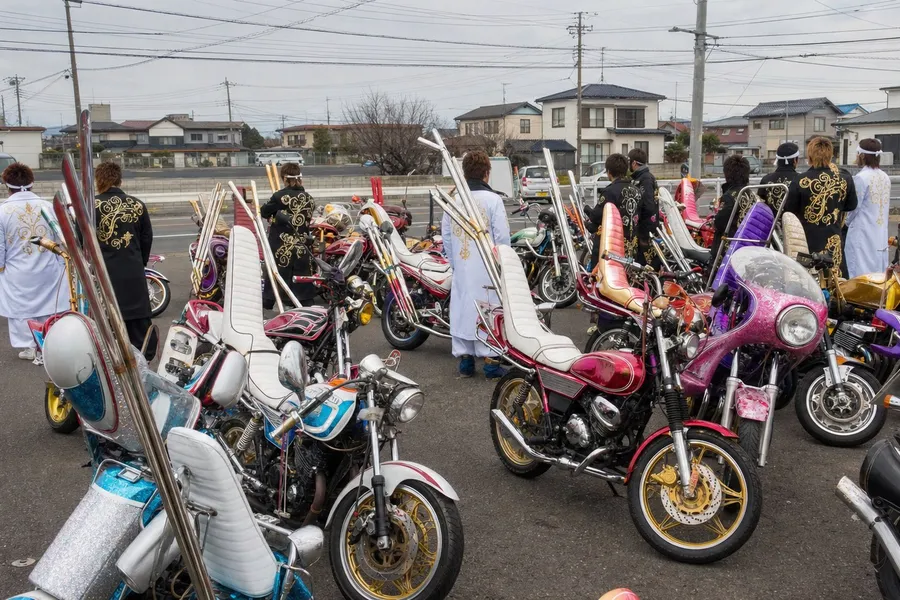 A photograph of a suburban parking area under a flat overcast sky, packed with about twenty extravagantly customised motorcycles parked at careless angles. Each machine differs from the others: towering three-tier stepped seats with tall padded backrests, narrow pull-back handlebars, bulbous rocket-shaped cowls and enormous chrome exhaust pipes sweeping up and back above the rear wheels, finished in candy metalflake pink, sky blue, purple, crimson, white and gold. Several young riders in long boxy white and black coats with bold gold embroidery stand among the machines, all seen from behind or with heads turned away so that no face is visible. Low houses and power lines run along the horizon. AI-generated photo illustration; not a photograph of any real gathering.