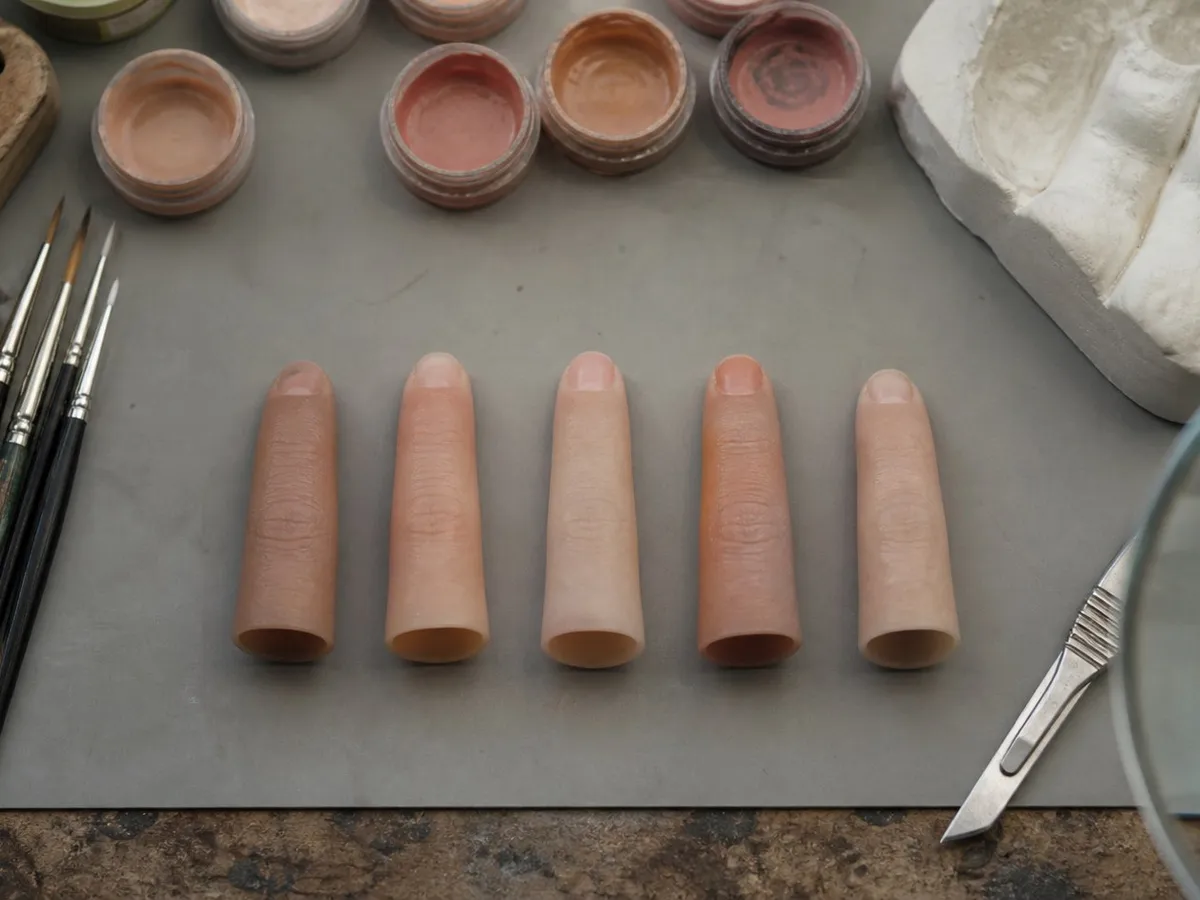 A photograph looking down on the workbench of a small prosthetics workshop. Five silicone prosthetic fingertips in slightly different skin tones lie in a row on a grey work mat, each hollow and open at its base. Around them are fine sable brushes, open pots of skin pigment, a scalpel, a plaster cast of a hand and the edge of a magnifying lamp. Everything is worn and orderly. No people and no hands appear. AI-generated photo illustration; not a photograph of any real workshop.