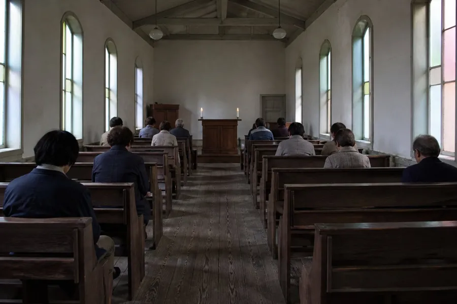 A photograph of the interior of a very small old Christian church in western Japan, taken from the back of the nave during a service. Ten or twelve people, most of them elderly, sit far apart on plain dark wooden pews, all seen from behind with heads slightly bowed; the great majority of the pews are empty. Whitewashed plaster walls, a worn plank floor, exposed ceiling beams, tall narrow windows of pale coloured glass, and a simple wooden table with two candles at the far end. Modest and poor but carefully kept. No faces are visible. AI-generated photo illustration; not a photograph of any real church, service or person.